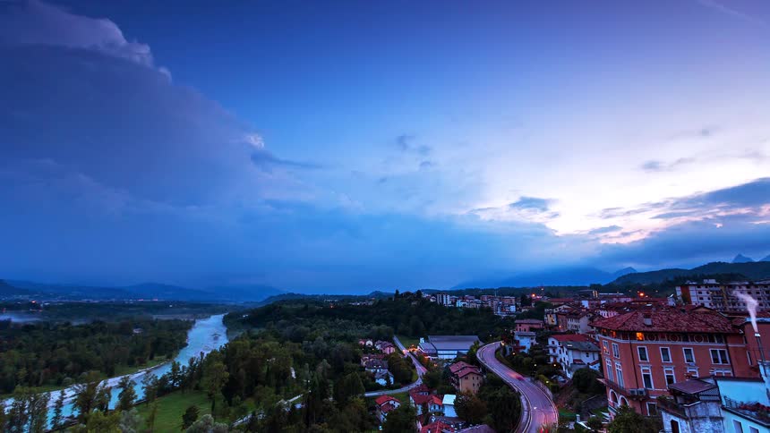 A wide view from above shows a winding river and a city as the sky turns from blue and pink to dark while city lights glow and car headlights move along a road