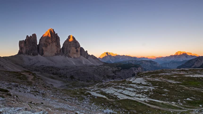 Sunlight illuminates rugged rocky mountain peaks rising above a vast alpine landscape under a clear blue sky