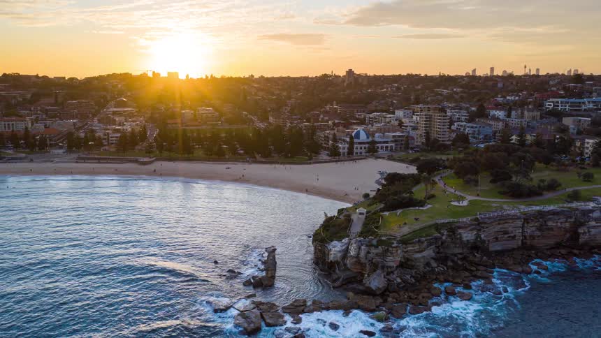 An aerial view shows a coastline with cliffs and a sandy beach next to a city as the sun sets over the horizon