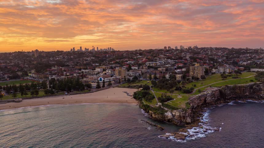 An aerial view shows a coastline with cliffs and a sandy beach next to a city as the sun sets over the horizon