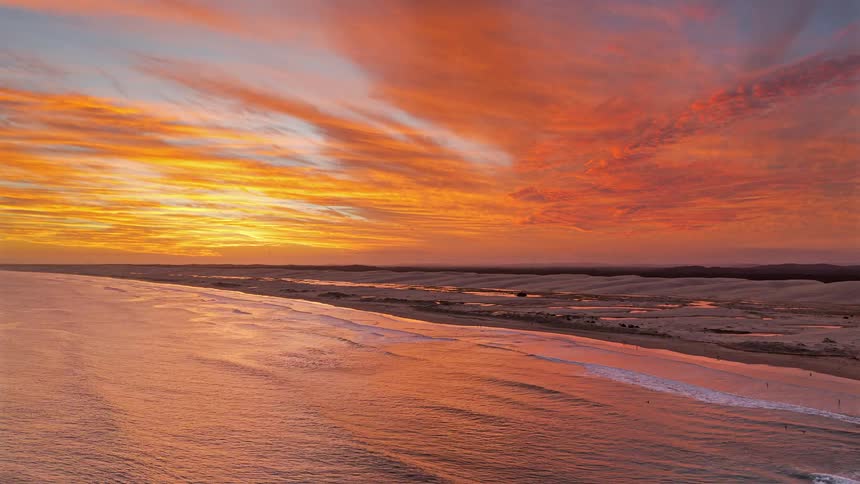 Golden sunset light spreads across ocean waves along a long sandy beach under dramatic colorful clouds