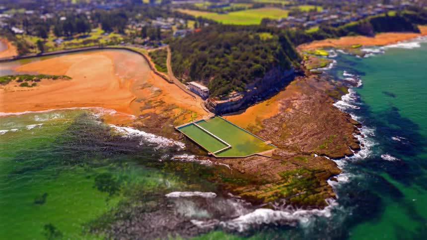 A rectangular ocean pool built into a rocky ledge between a golden sand beach and the turquoise sea while green hills and coastal houses sit in the background under bright daylight