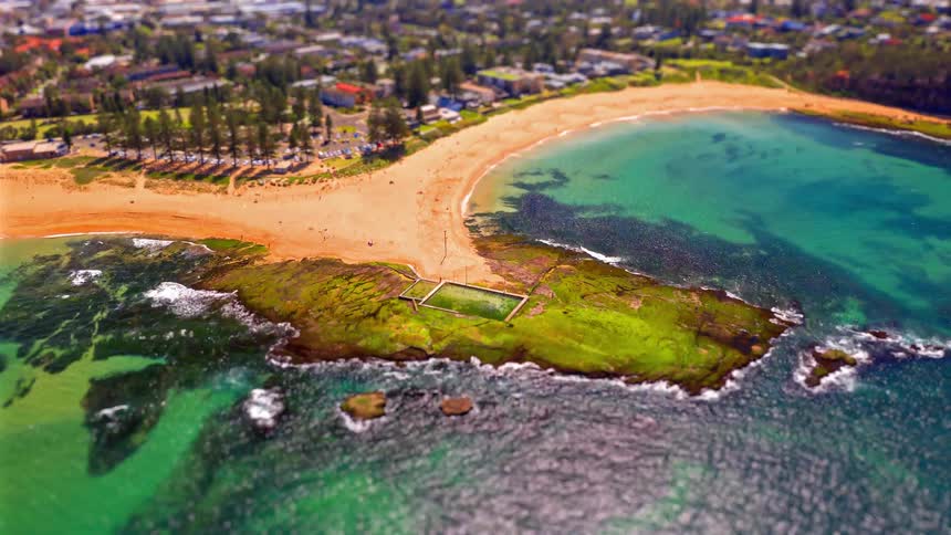 An aerial view shows a curved sandy beach and turquoise water surrounding a small green peninsula near a coastal town