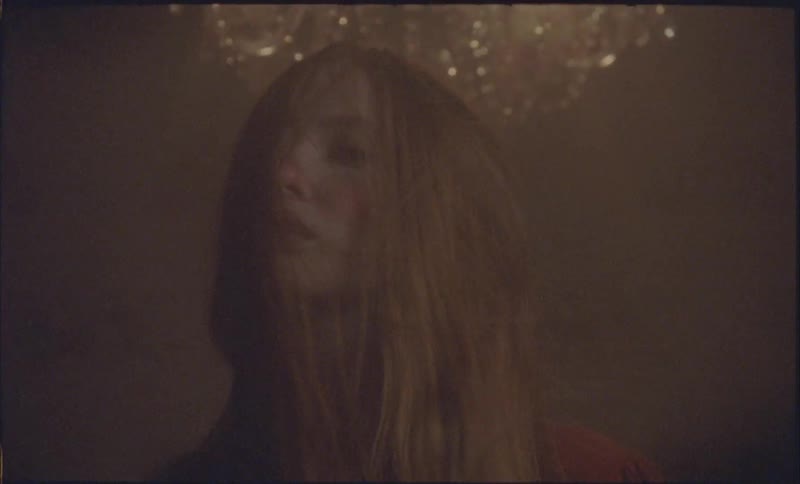 A blonde woman with long hair stares to the camera with a confident expression under a glowing crystal chandelier in a dark room
