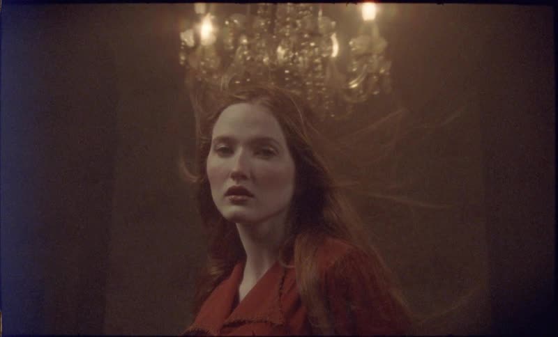 A restless wind moves through the dim room lifting the womans hair as the chandeliers lights tremble softly above her