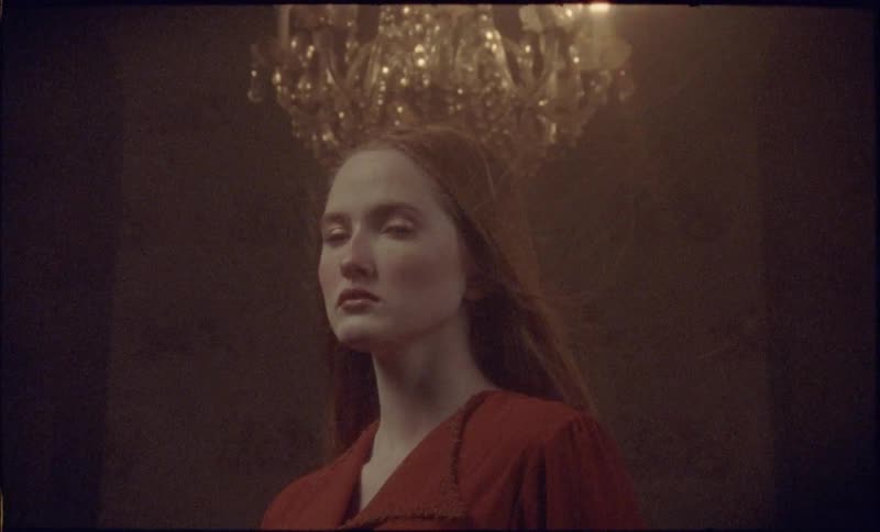 A closeup of a woman in a red outfit standing beneath a glowing chandelier in a windy room her face calm and serious in the dim light