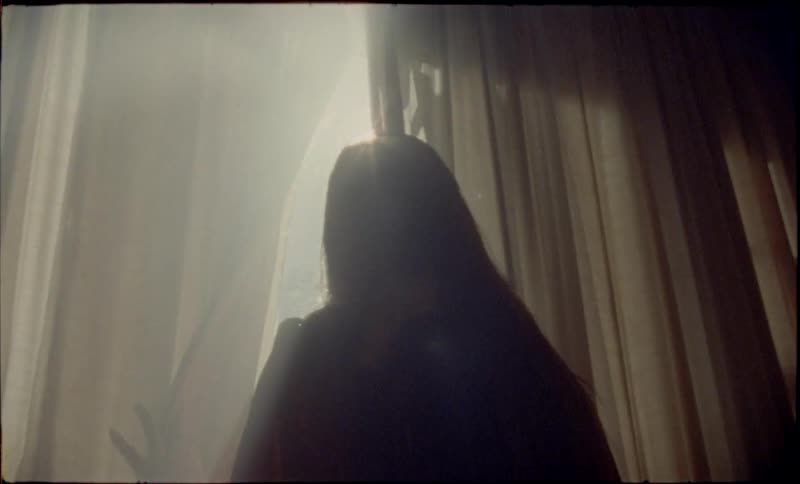 A view from behind shows a woman standing in a dark room as she opens the curtains to let bright sunlight inside
