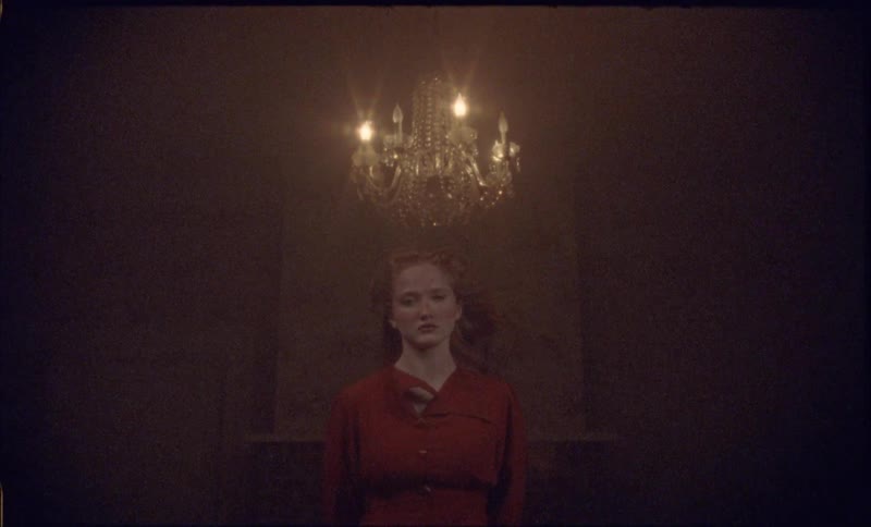 A view shows a girl with long red hair wearing a red dress standing in a dark room under a glowing glass chandelier