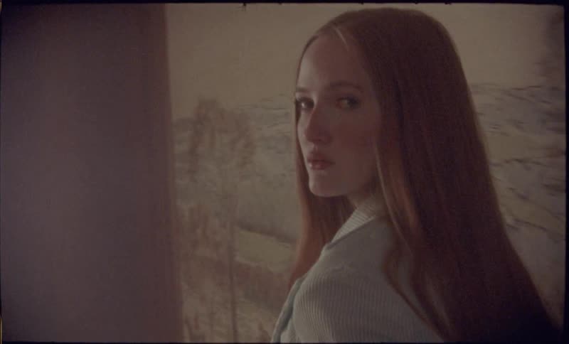 A young woman with long ginger hair turns her head to look directly at the camera while standing in a room next to a large painting of a winter village scene
