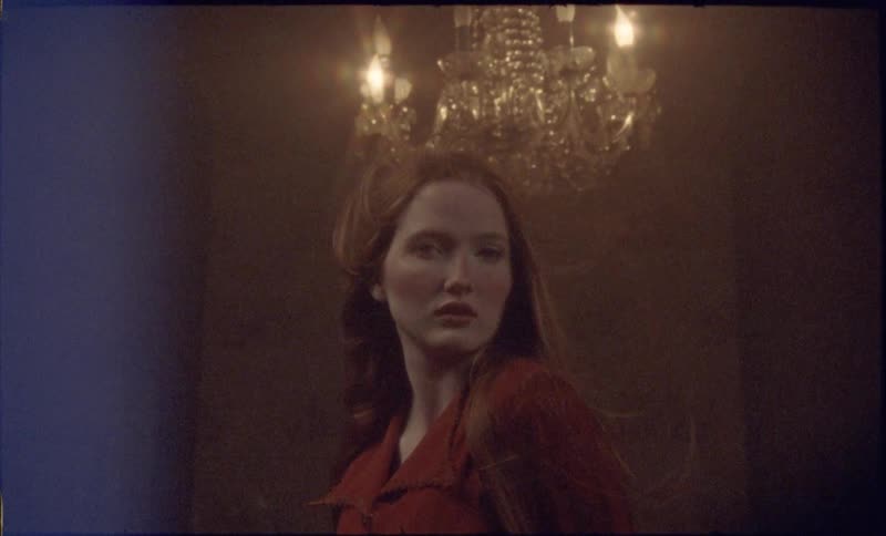 A woman with long red hair stands in a dimly lit room beneath a glowing crystal chandelier