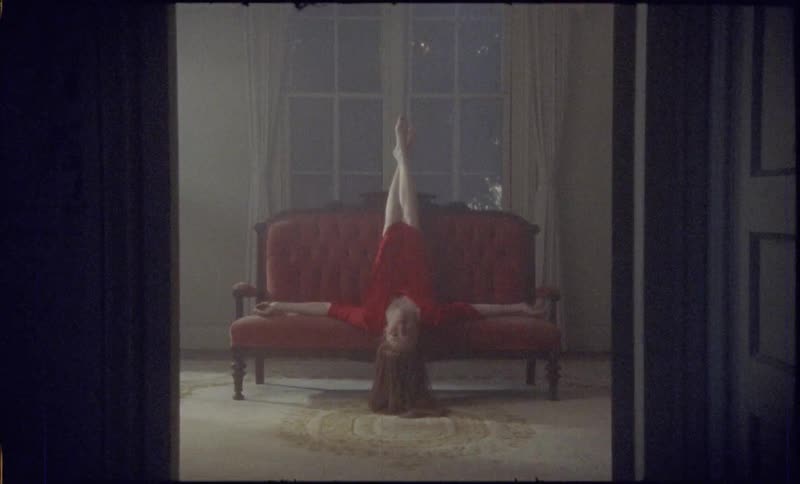 Young woman lies with her legs in the air and her head hanging off the love seat in a dimly lit room
