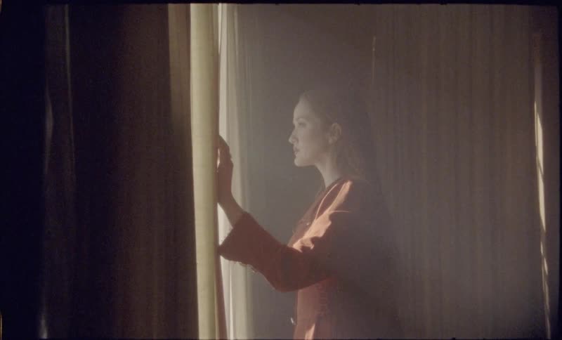 Woman holds a curtain while looking through a lit window