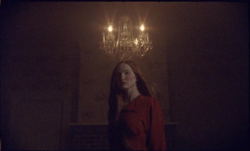 A woman in a red dress stands beneath a glowing chandelier in a dim windy moody room gazing forward with a calm but mysterious expression