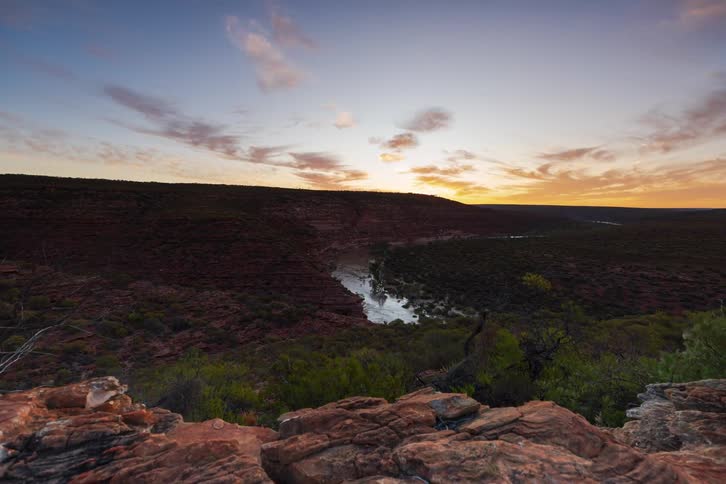 A view from a rocky cliff shows a river winding through a valley as the sun sets and the sky changes from blue to orange and purple