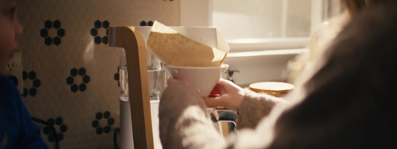 Woman puts a filter on a coffee pot as her young son sits on the counter watching