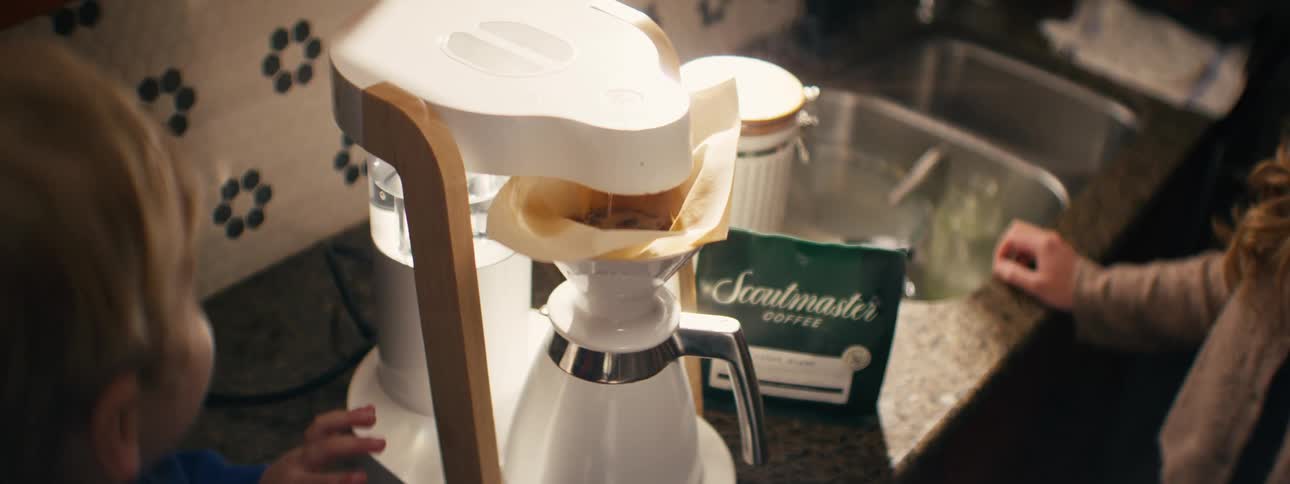 A young child touches the paper filter already placed inside the coffee maker while a bag of scoutmaster coffee sits on the counter nearby