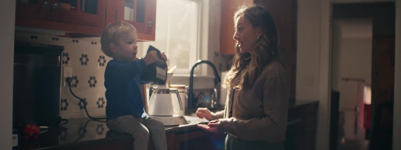 Woman standing in the kitchen with her little boy sitting on the counter as she hands him a bag of coffee and he shakes it