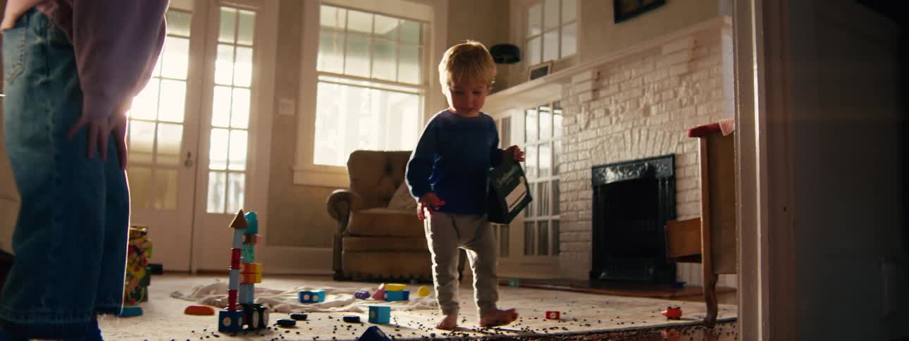 Small round objects are scattered across a wooden floor as a child walks barefoot nearby while toys sit on a rug in the background