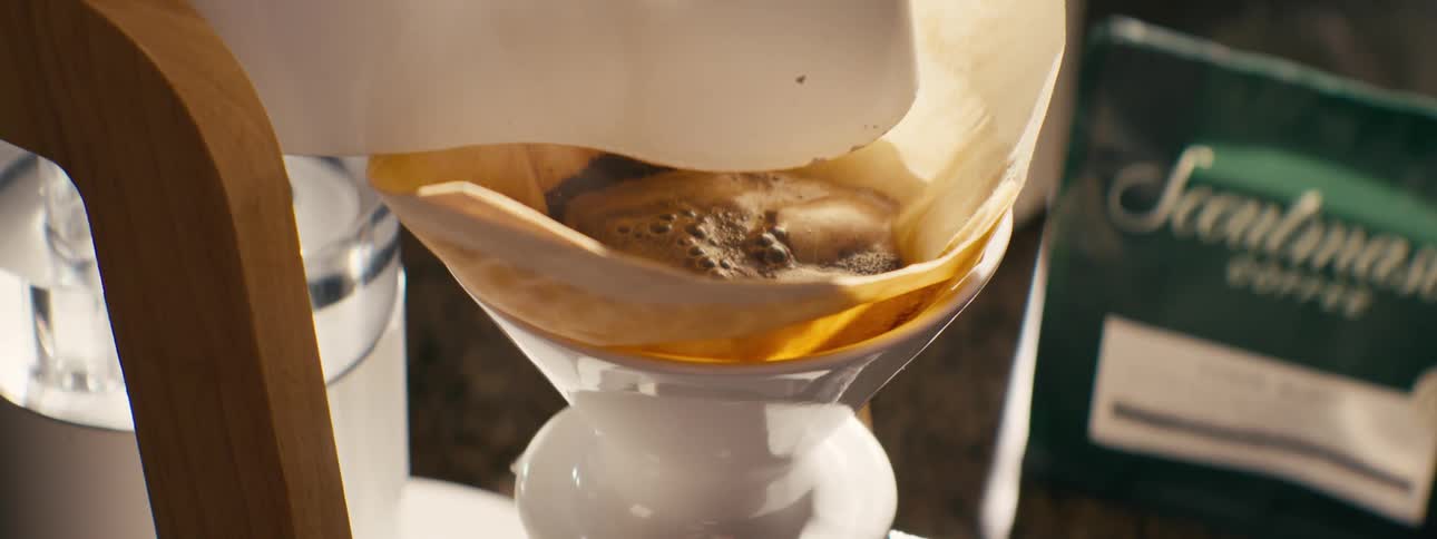 A close view shows water dripping into a paper filter filled with coffee grounds as it brews inside a white coffee maker