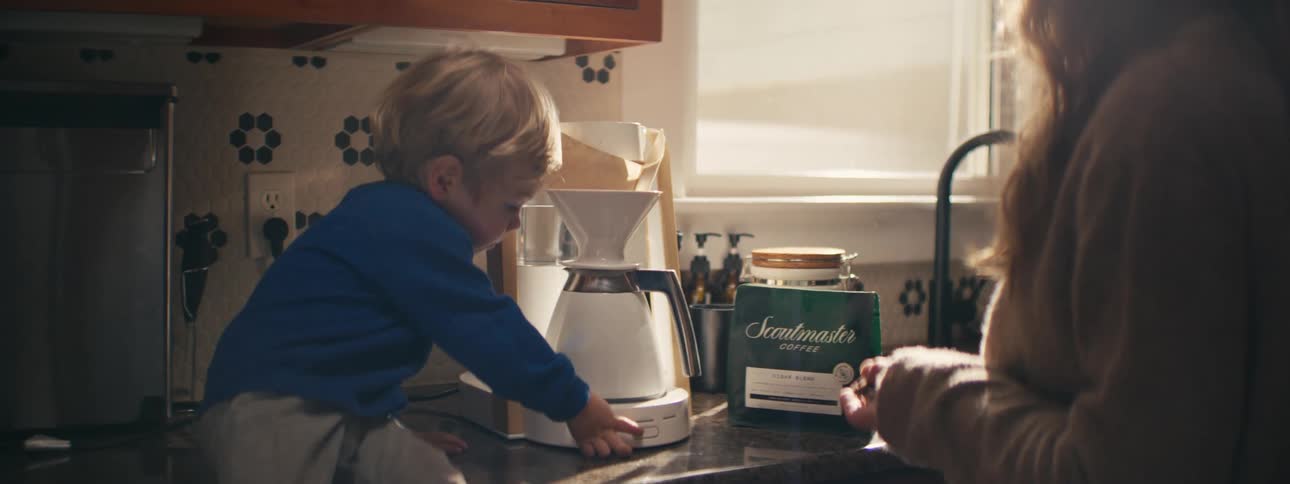 A young boy in a blue shirt reaches for a white electric pour over coffee maker on a granite counter while a woman holds a bag of coffee beans in a sunlit kitchen with a tiled backsplash