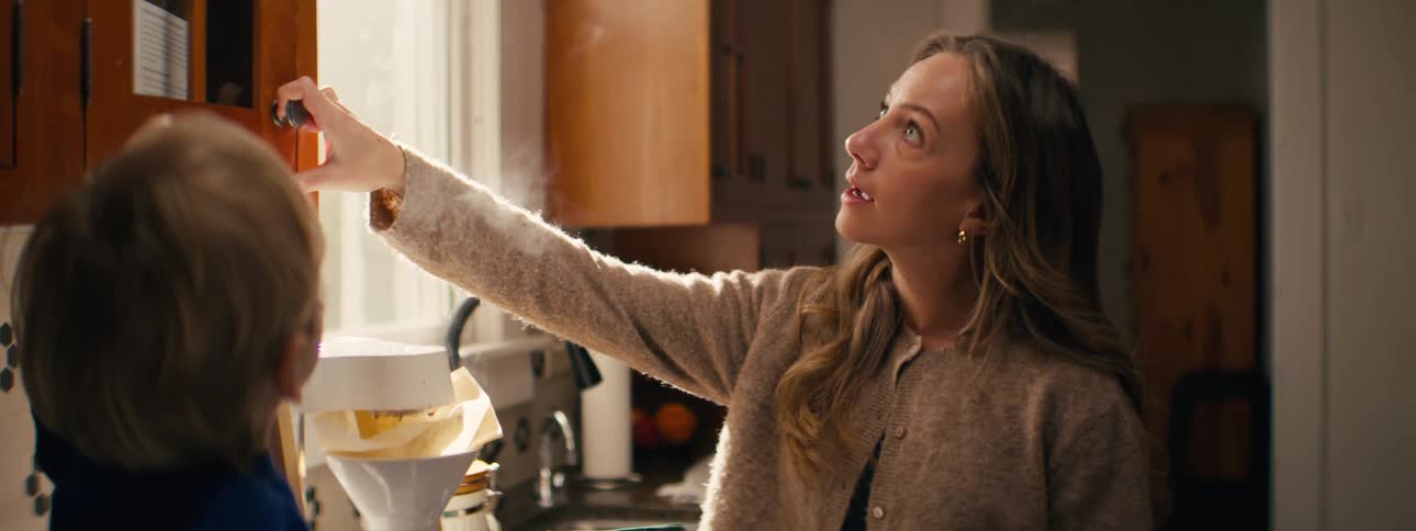 A woman reaches up to open a wooden cabinet in a bright kitchen while steam rises from a coffee maker on the counter below her