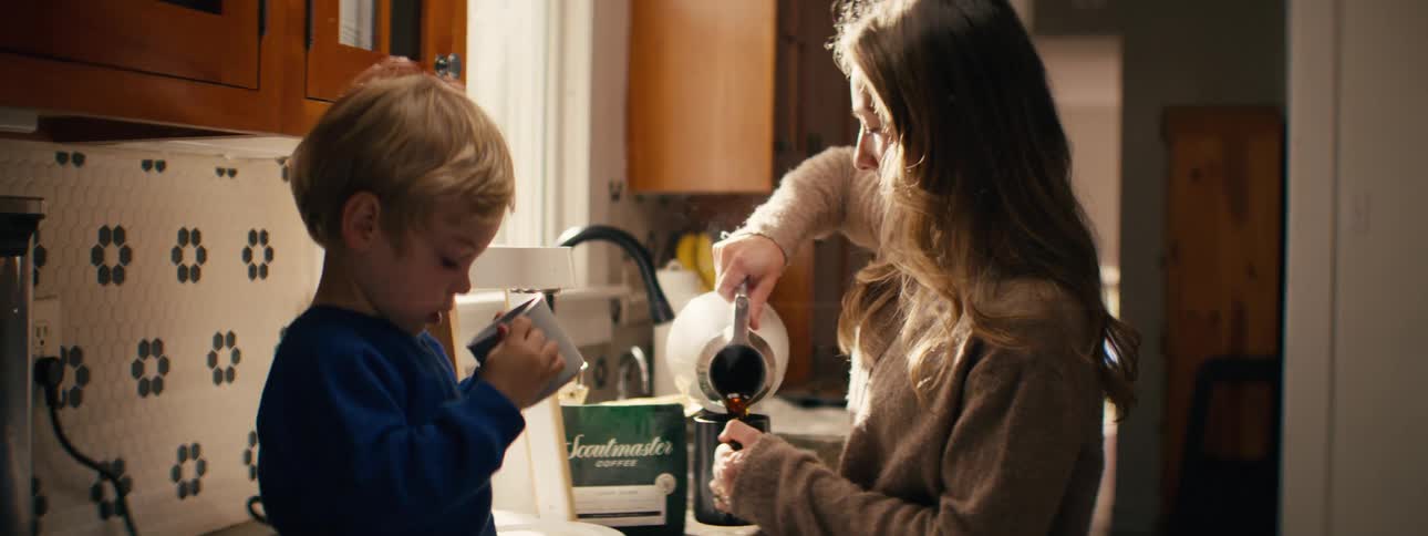 Woman pours coffee into a cup while a child holds another in the kitchen