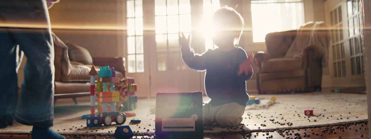 A young girl in a pink sweatshirt joins a blonde toddler boy on a sunlit carpet to play with a dark bag of coffee and hundreds of scattered coffee beans near a colorful toy tower