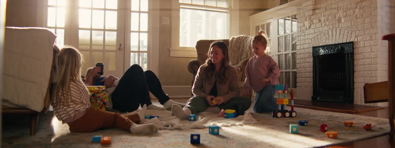 A family with three young children plays with building blocks on a rug in a sunlit living room as the father joins them and holds a mug
