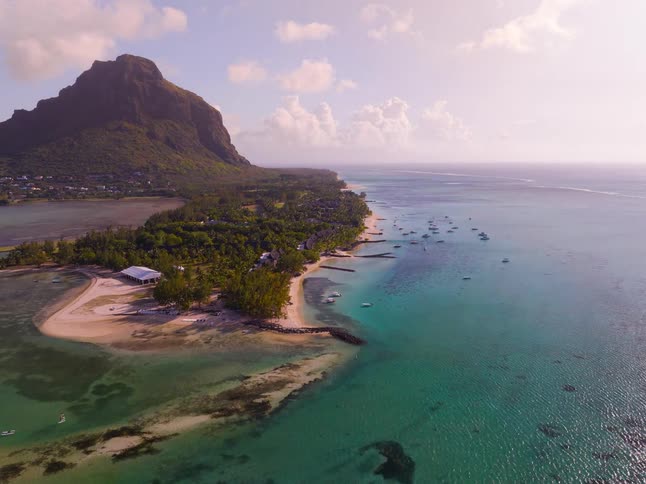 The video shows a highaltitude aerial sweep of a tropical coastline featuring clear turquoise water and a white sand beach lined with palm treesa massive rugged mountain peak dominates the background casting a long shadow over the lush