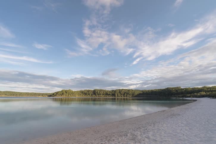 Forested mountains along the coast with people walking on the beach as clouds move across the sky and reflect on the water