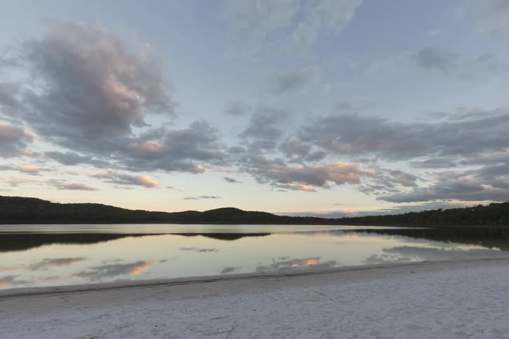 Forested mountains along the coast with a bird moving across the water near the beach as clouds move across the sky and reflect on the water