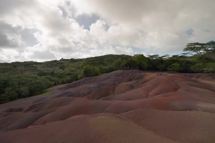 The seven coloured earths in mauritius featuring rolling sand dunes with distinct layers of red brown and purple sedimentthe landscape is surrounded by lush green tropical forests under a bright cloudy sky with a viewing platform visible in the bac