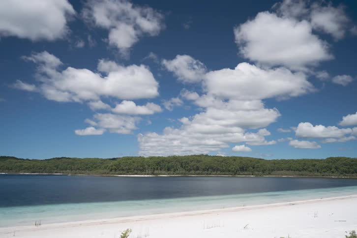 A calm lake with a sandy shoreline and forested hills under a blue sky with scattered clouds
