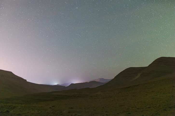 Mountains across the desert landscape as cloud move across the sky and the sun sets with stars glowing and traffic moving through the mountains