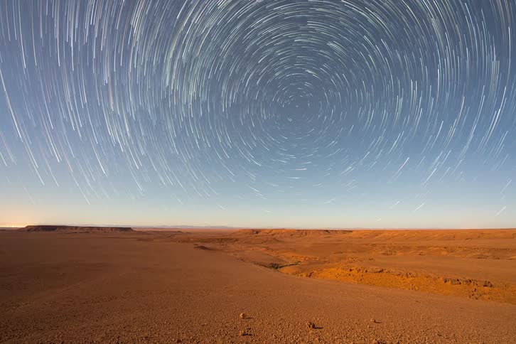 Long exposure star trails spinning in circular patterns over a barren desert landscape at night