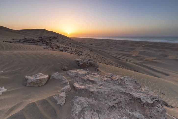 The sun sets over a desert landscape where sand dunes meet the ocean shoreline