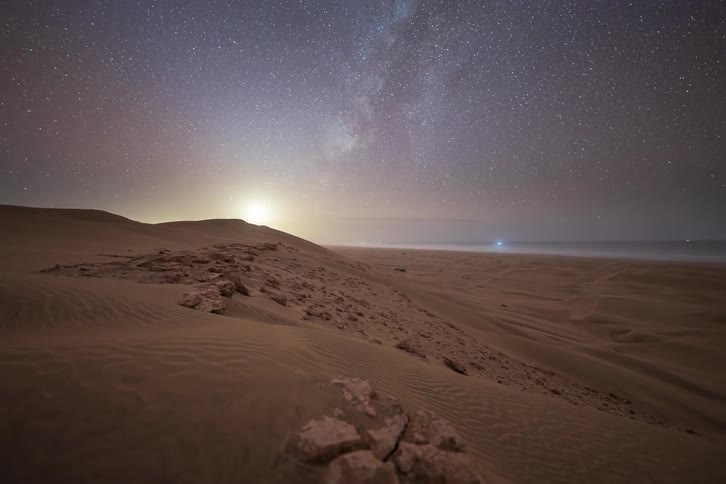 Mountains along the coast as boats sail on the water and the stars of the milky way glow in the sky as the sun sets