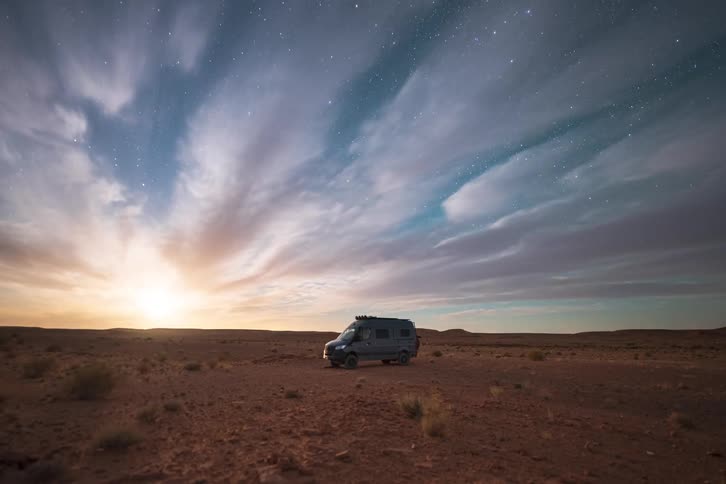 A grey camper van sits parked in the middle of a vast desert under a clear night sky filled with stars and glowing light on the horizon as thin clouds drift across the dark blue atmosphere above the rocky ground