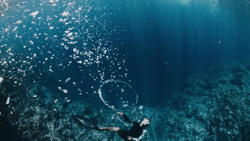 A freediver in a black wetsuit and fins swims through a deep blue underwater cavern while a large trail of bubbles and a single bubble ring float toward the sunlit surface near a dark rocky reef