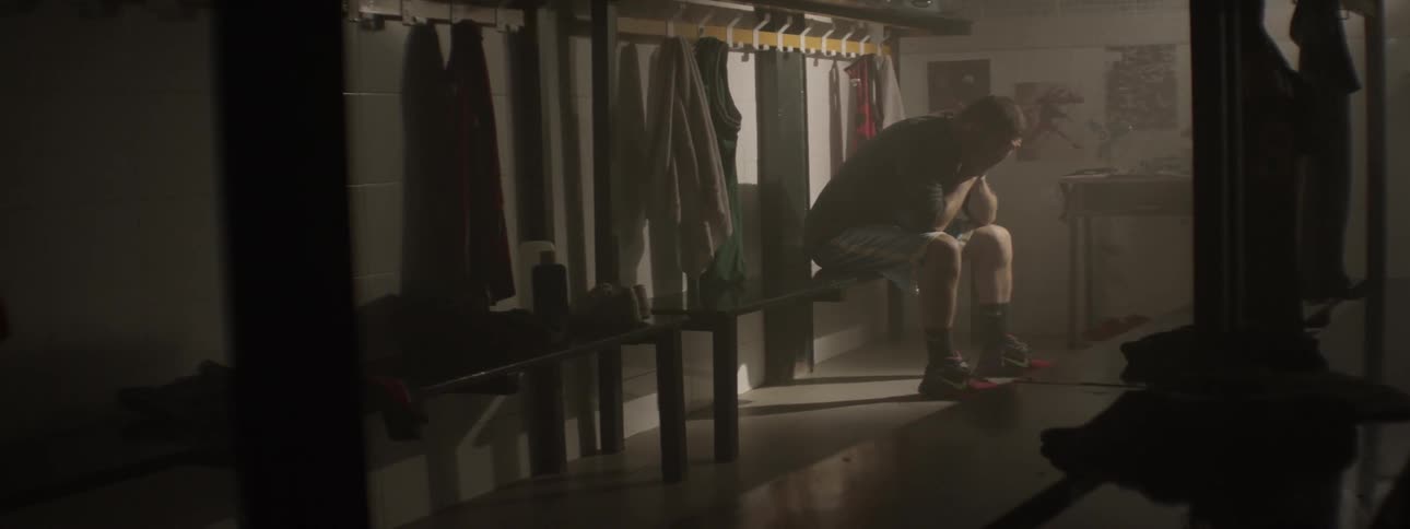 A man sits in a dark locker and ties his shoes