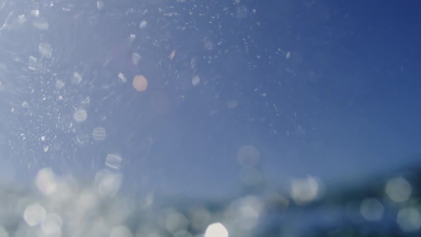 The camera starts above the ocean surface catching bright sunlight and then quickly plunges underwater where a dense cloud of white bubbles swirls against a deep blue background as light rays shimmer through the moving water