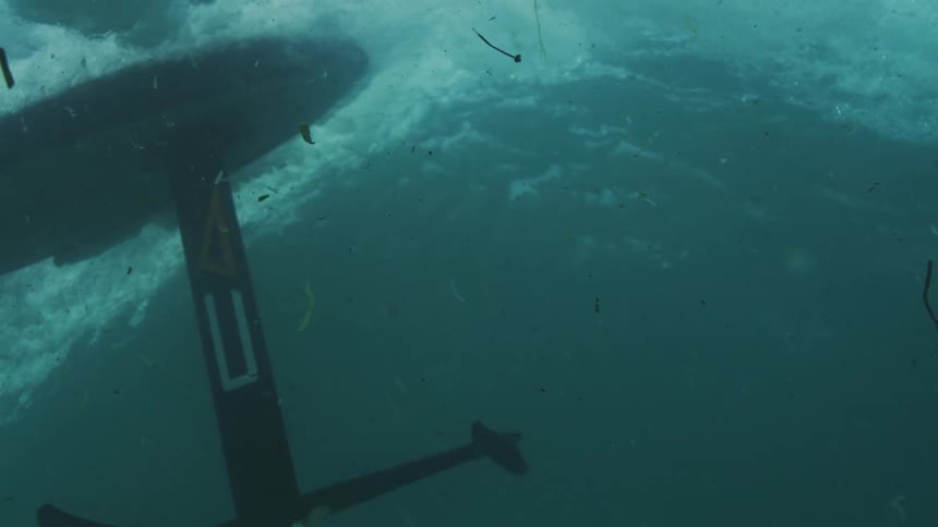 Underwater footage shows the carbon fiber foil and mast of a hydrofoil surfboard slicing through dark debrisfilled wateras a wave breaks above the board is engulfed by a chaotic wall of white bubbles and turbulent foam