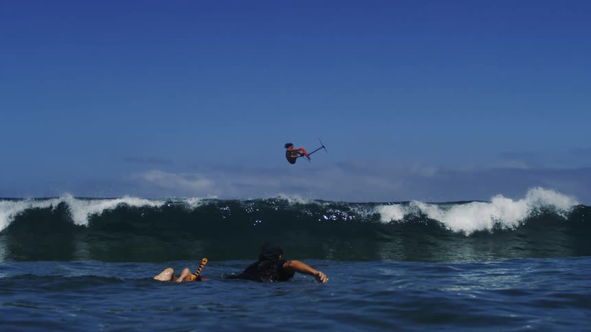 Surfer leaps intot he air with his board while another surfer lies on his board on a summer day