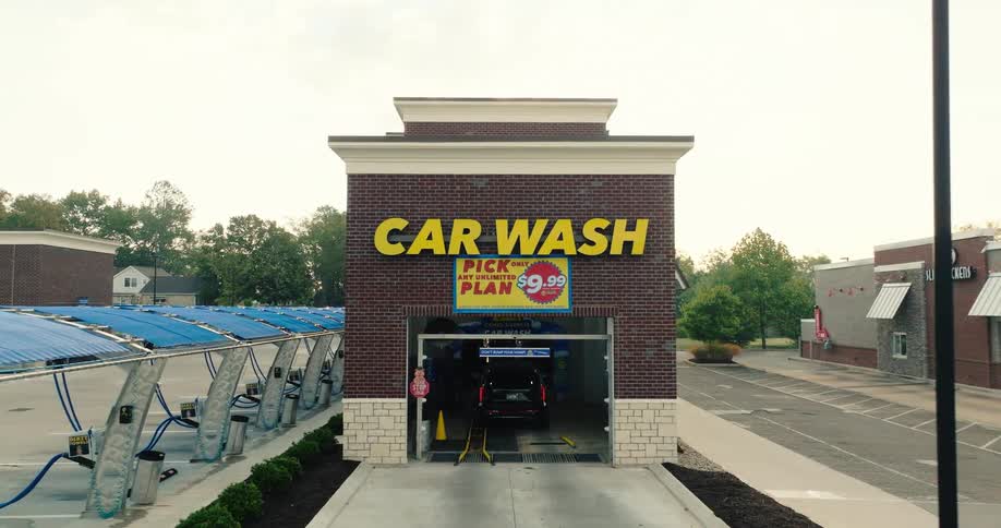 The video features a brick car wash building with a large yellow car wash sign and a promotional banner for a 999 unlimited plan to the left of the main bay there is a row of vacuum stations under blue awnings while a car is seen entering the wash