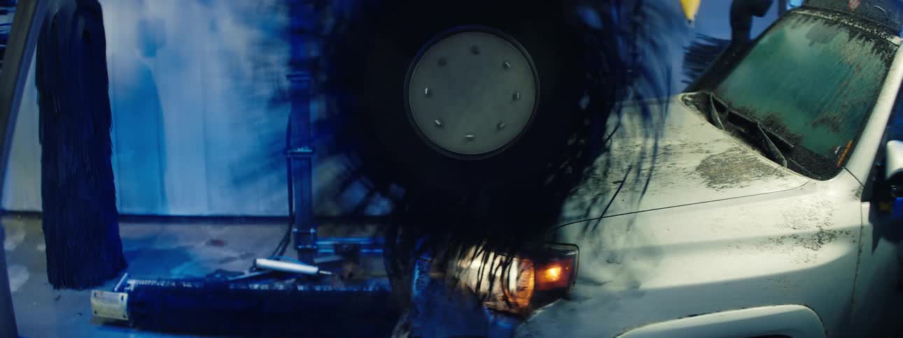 A dirty white suv slowly drives through an automated car wash while a large black rotating brush cleans the hood and side of the vehicle against a blue wall