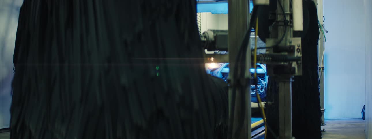 Through a gap between large rotating black carwash brushes and metal machinery a blue car is partially visible inside an automated wash bay illuminated by cool industrial lighting
