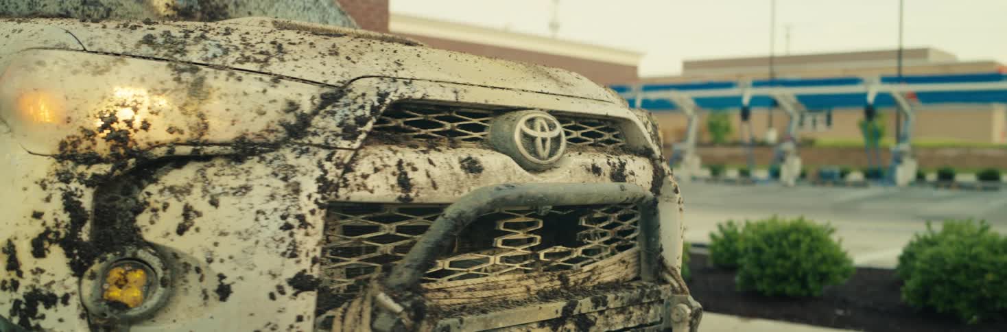 The video shows a closeup of a white toyota 4runner heavily caked in dark thick mud across its front grille and headlightsas the camera pans around the vehicle the setting is revealed to be a car wash parking lot with a brick building i
