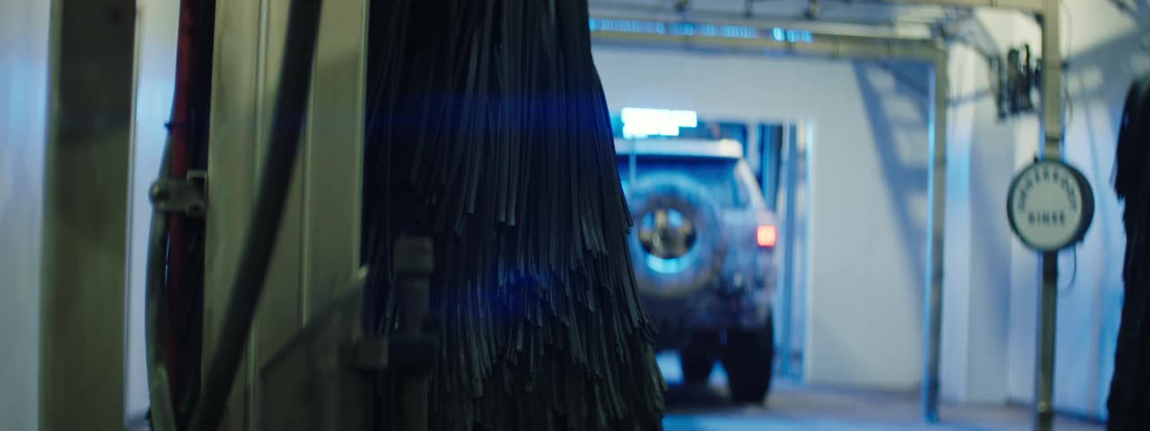 A large offroad vehicle with a spare tire on the back exits an automated car wash thick black hanging brushes frame the foreground as the tan suv pulls away into the bright garage