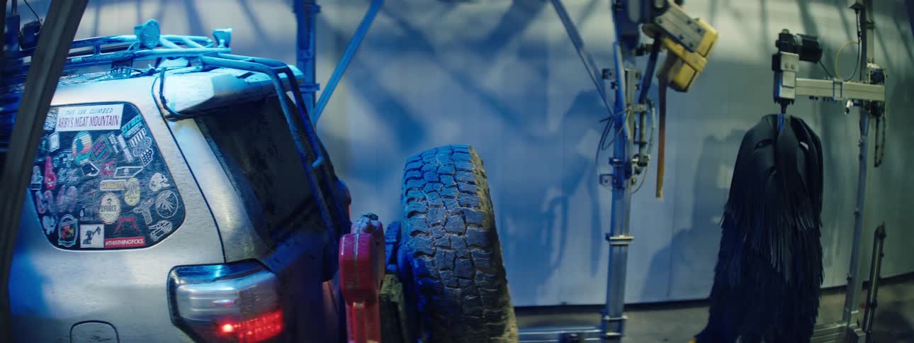 A mudstreaked suv adorned with a collage of colorful stickers on its rear window glides through a car wash tunnel where cool blue light highlights the dirt as the vehicle passes beneath the industrial machinery
