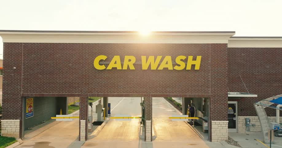 Golden sunlight crests over the large yellow sign of a modern brick car wash casting long shadows across the empty pavement as the entrance lanes wait under the warm hazy glow of late afternoon